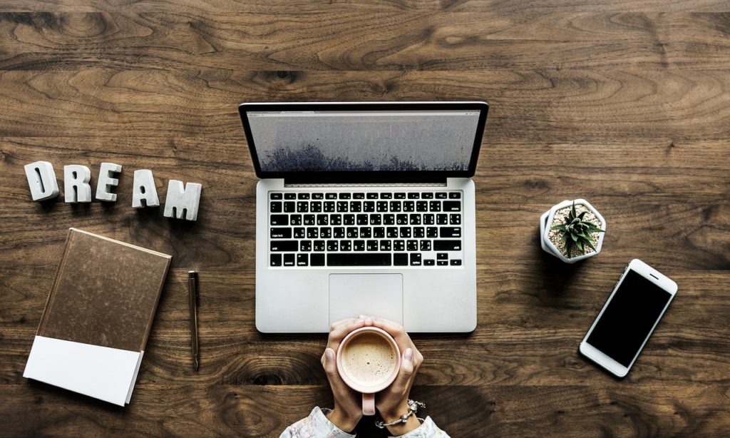 Desktop with a laptop and someones hands holding a cup of coffee.  Also on the desktop is the word DREAM, a notebook, cell phone and small plant.