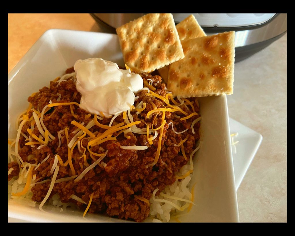 A white square bowl filled with no bean chili topped with shredded cheese, sour cream and saltine crackers.