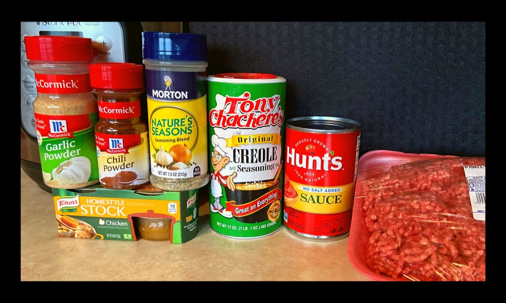 A instant pot, chili powder, chicken broth, nature seasoning, tony chachere's seasoning, can of tomato sauce, and raw ground meat all sitting on a counter top.