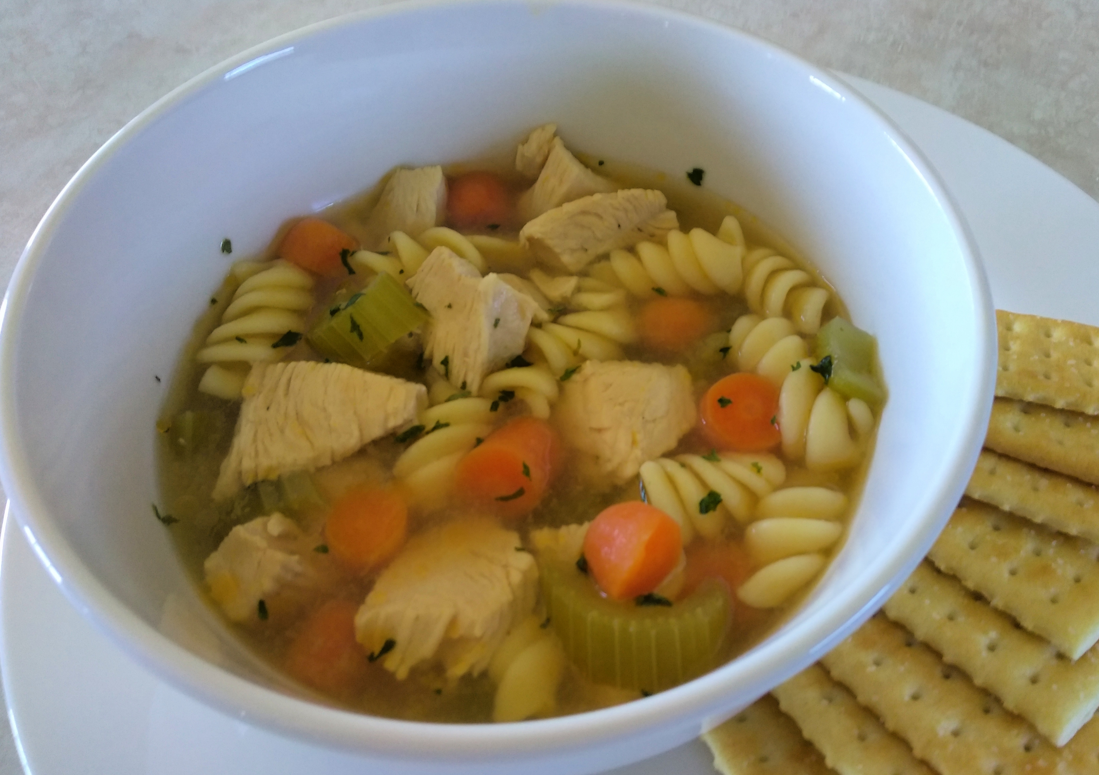 White bowl on a counter top filled with chicken noodle soup with crackers on the side.