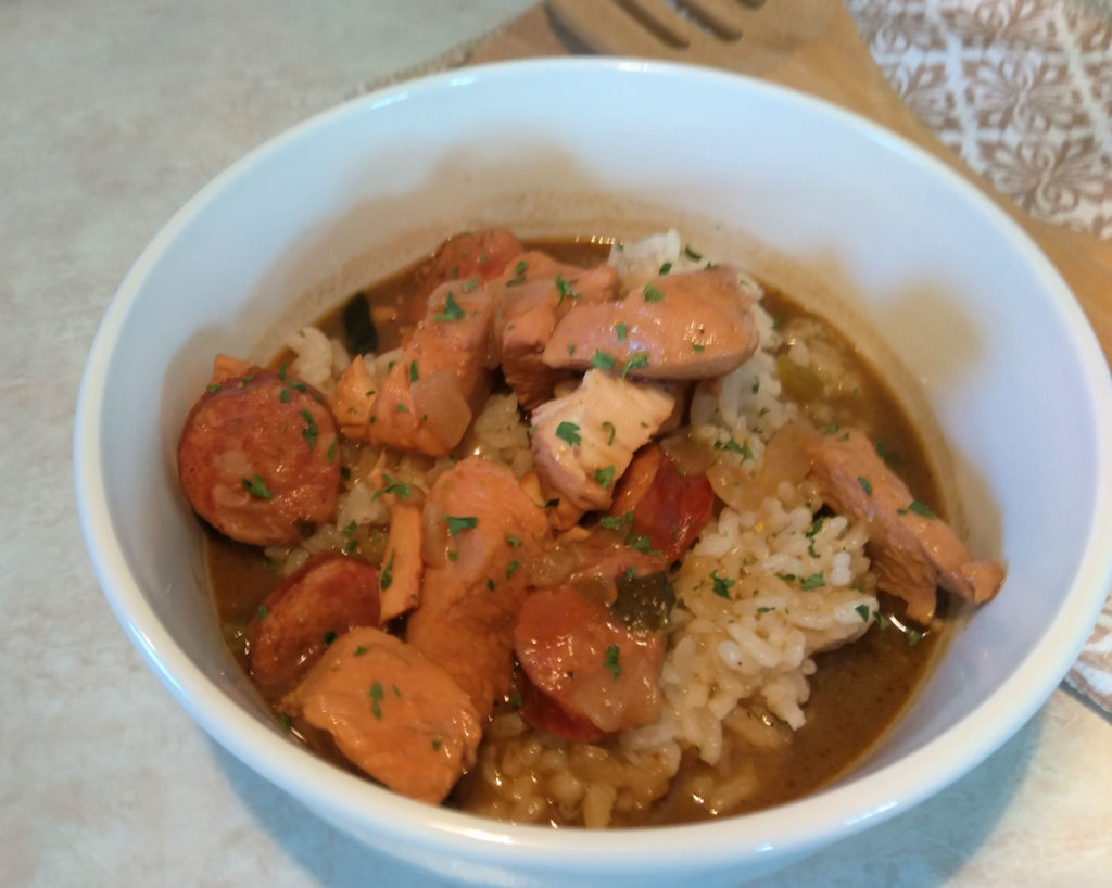 A white bowl of chicken and sausage gumbo on a counter top. A wooden spoon and decorative napkin in the background.