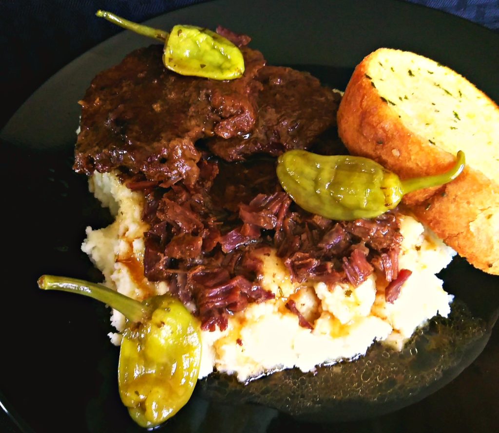 A black plate with Mississippi Pot Roast and gravy over mashed potatoes with a garlic bread.