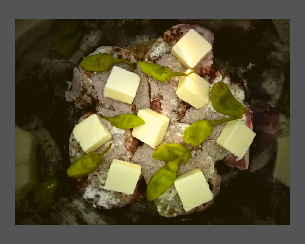 A silver Instant Pot filled with chuncks of pork roast with packets of ranch and au jus sprinkled on top. A sliced stick of butter on top with peperoncini peppers.