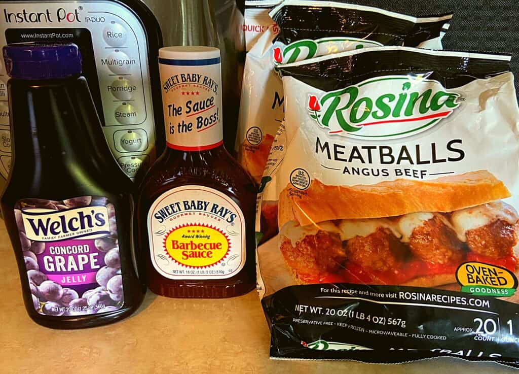A bottle of welchs grape jelly, bottle of sweet baby rays bbq sauce, and two bags of frozen meatballs.