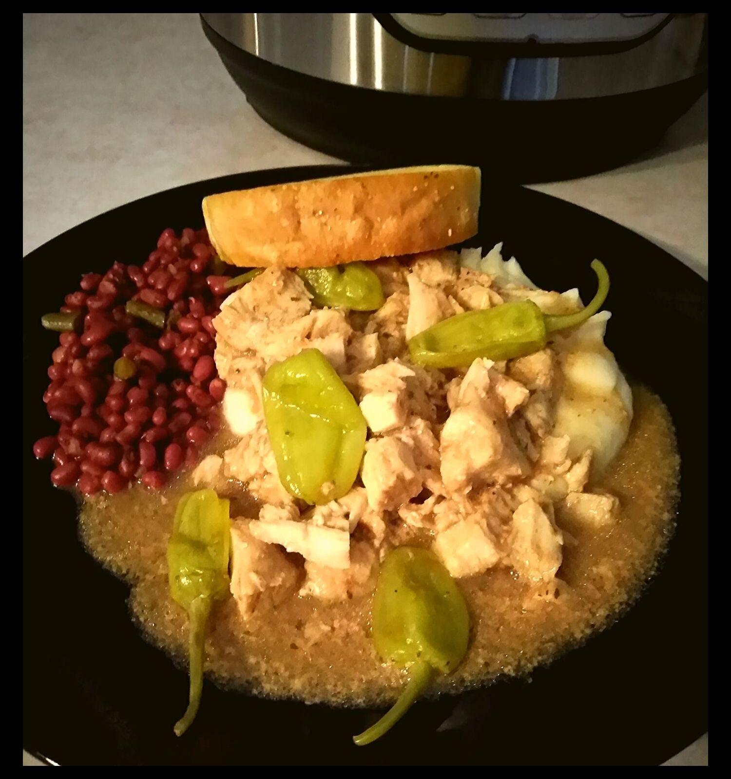 A black plate filled with mississippi chicken over mashed potatoes with field peas and a garlic bread.