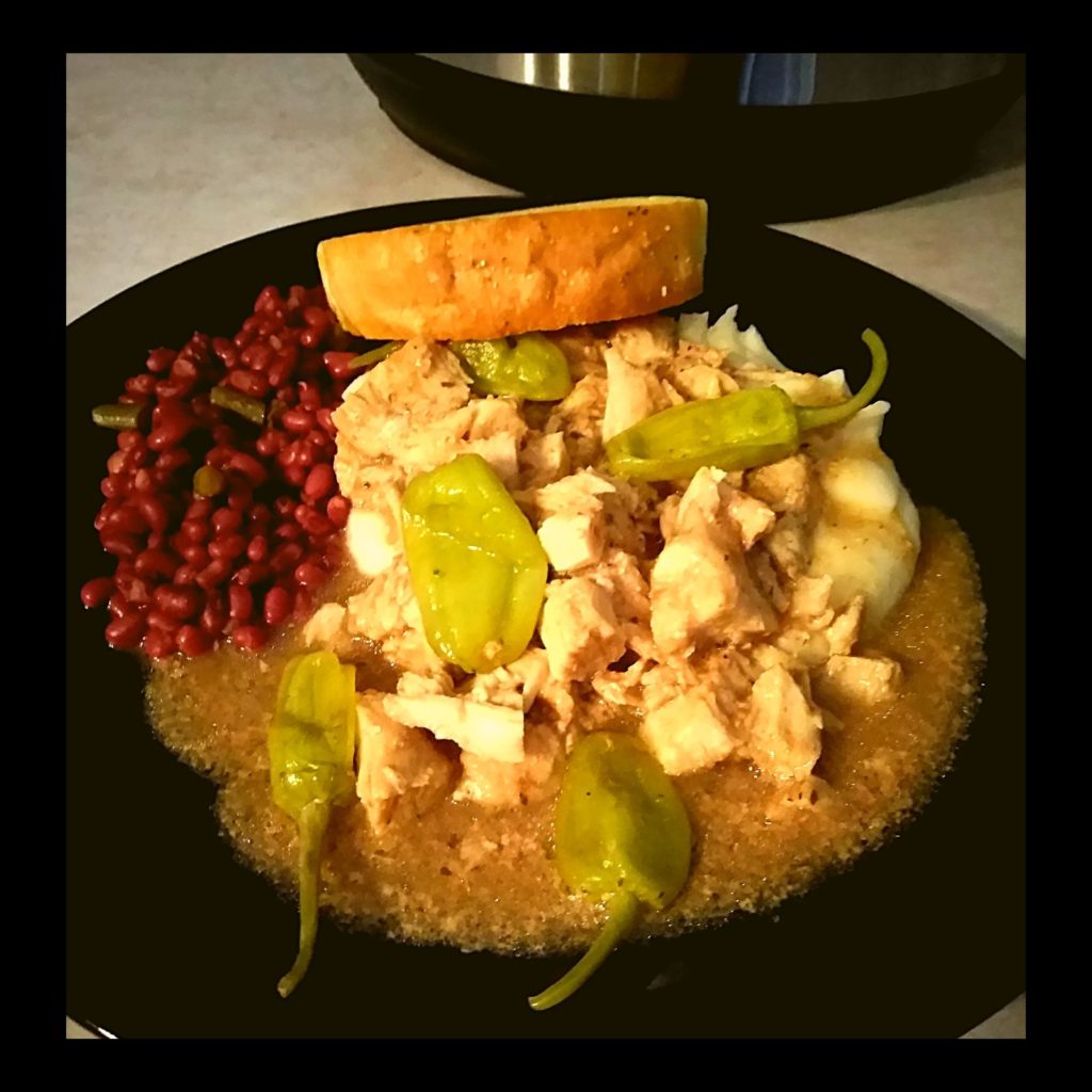 A black plate filled with mashed potatoes and gravy topped with mississippi chicken, pepperoncini peppers and field peas.
