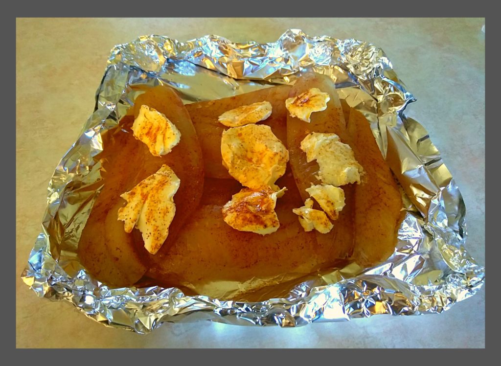Raw tilapia fillets in a foil style boat with seasoning and butter on top.