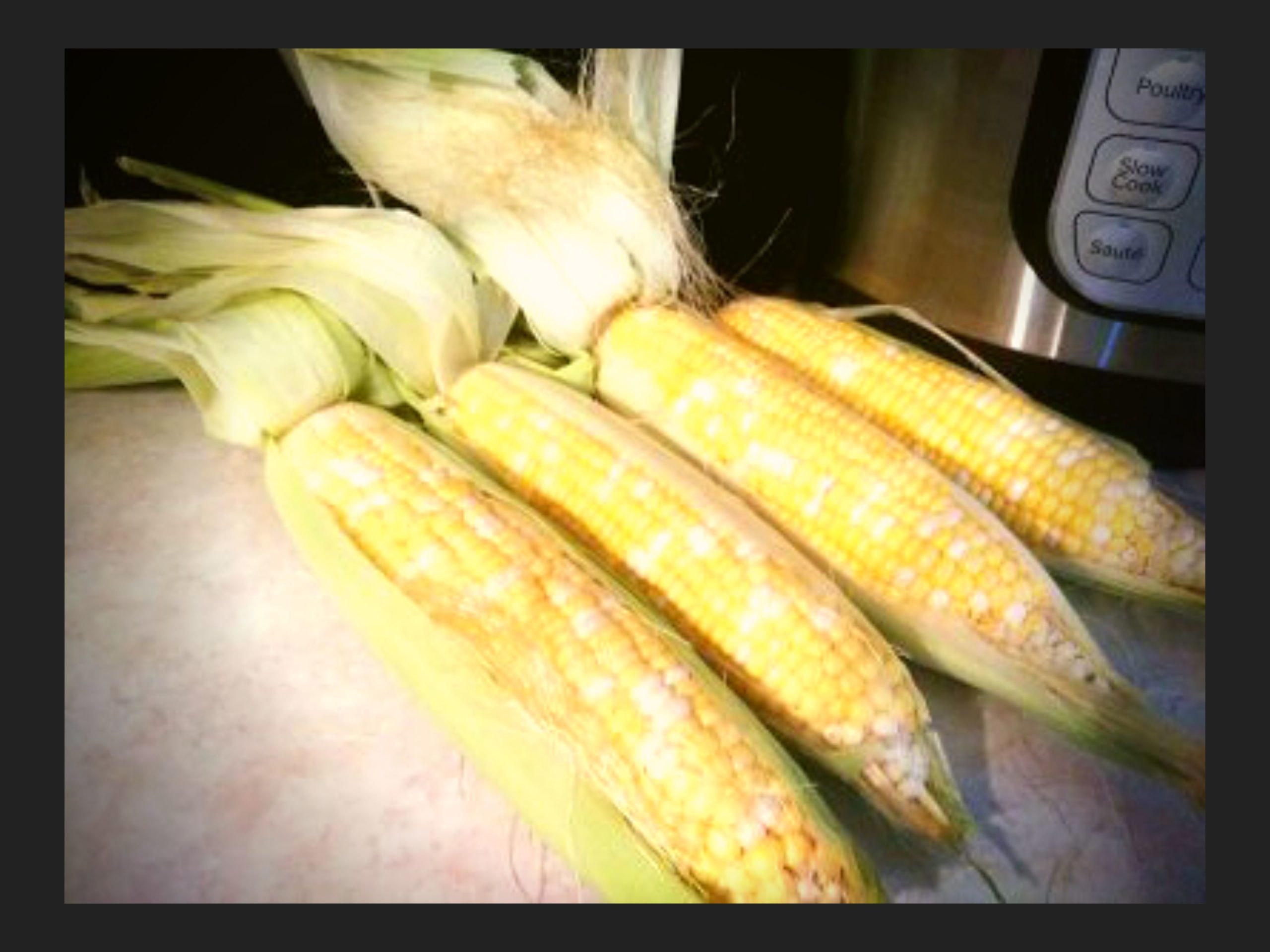 4 ears of corn with the husk pulled half way off on a counter top next to an Instant Pot.