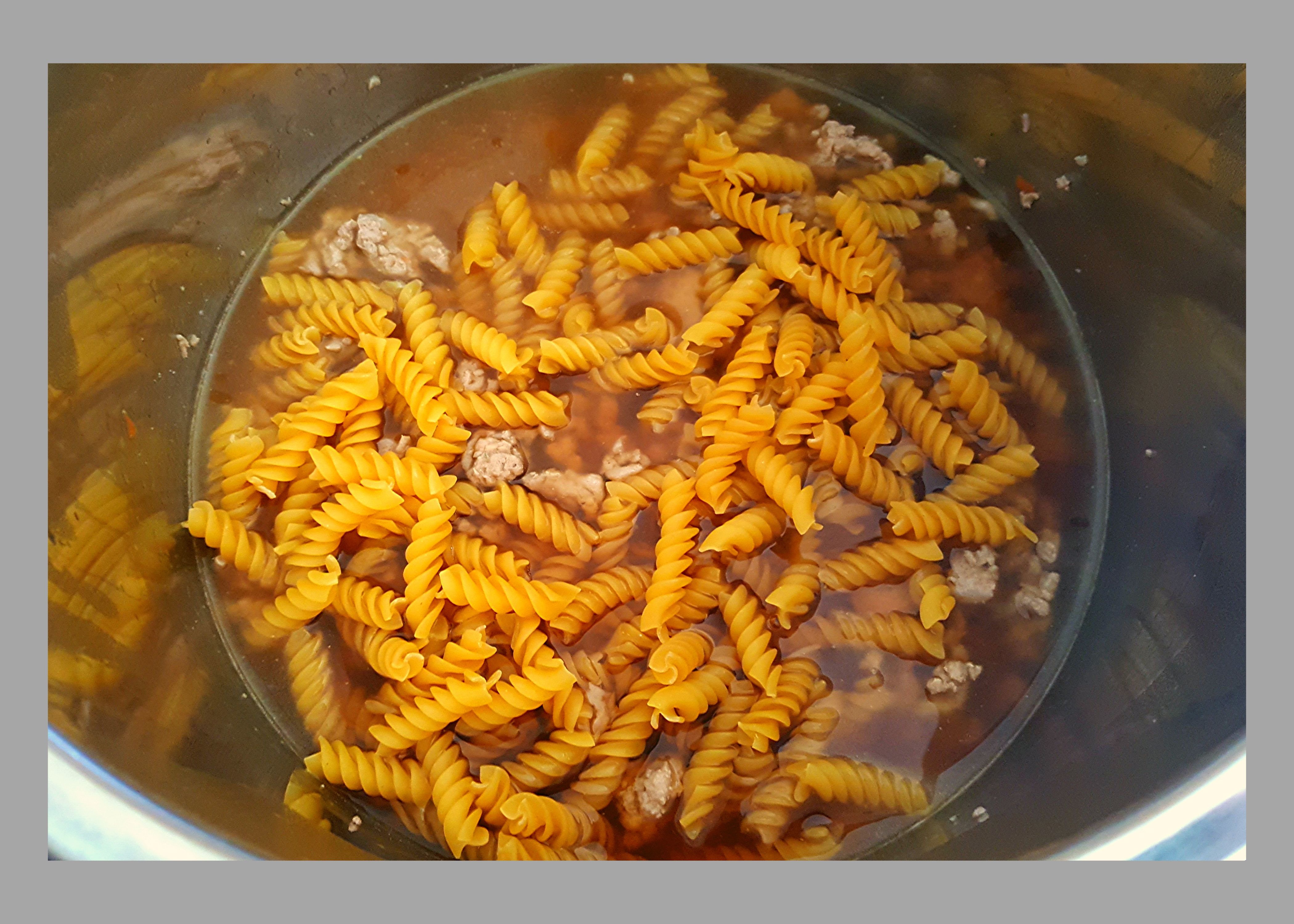 The inside of a silver Instant Pot filled with ground turkey, beef broth, and uncooked spiral noodles.