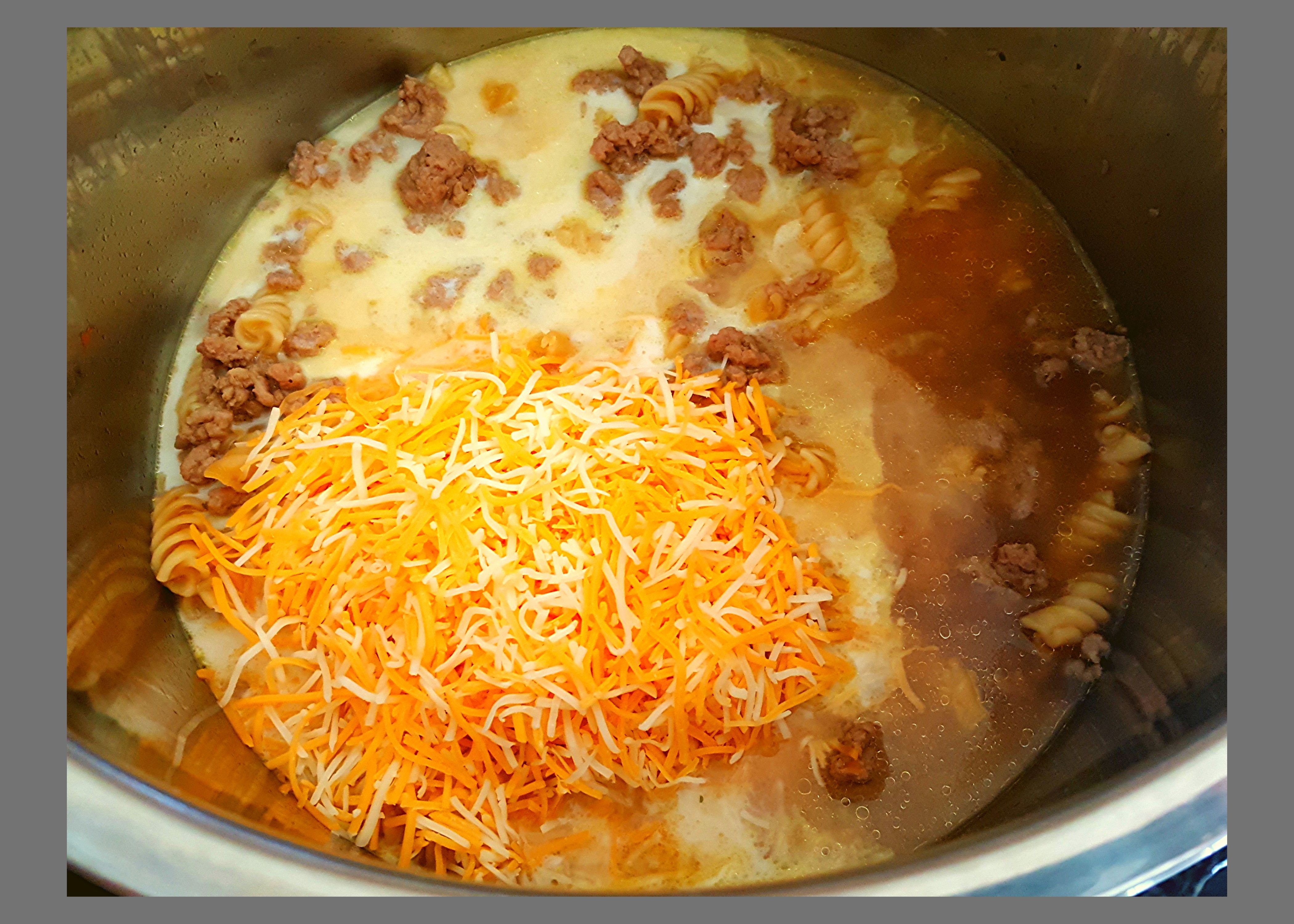The inside of an Instant Pot filled with a cup of shredded cheese on top of cooked ground turkey and spiral noodles that needs to be stirred.