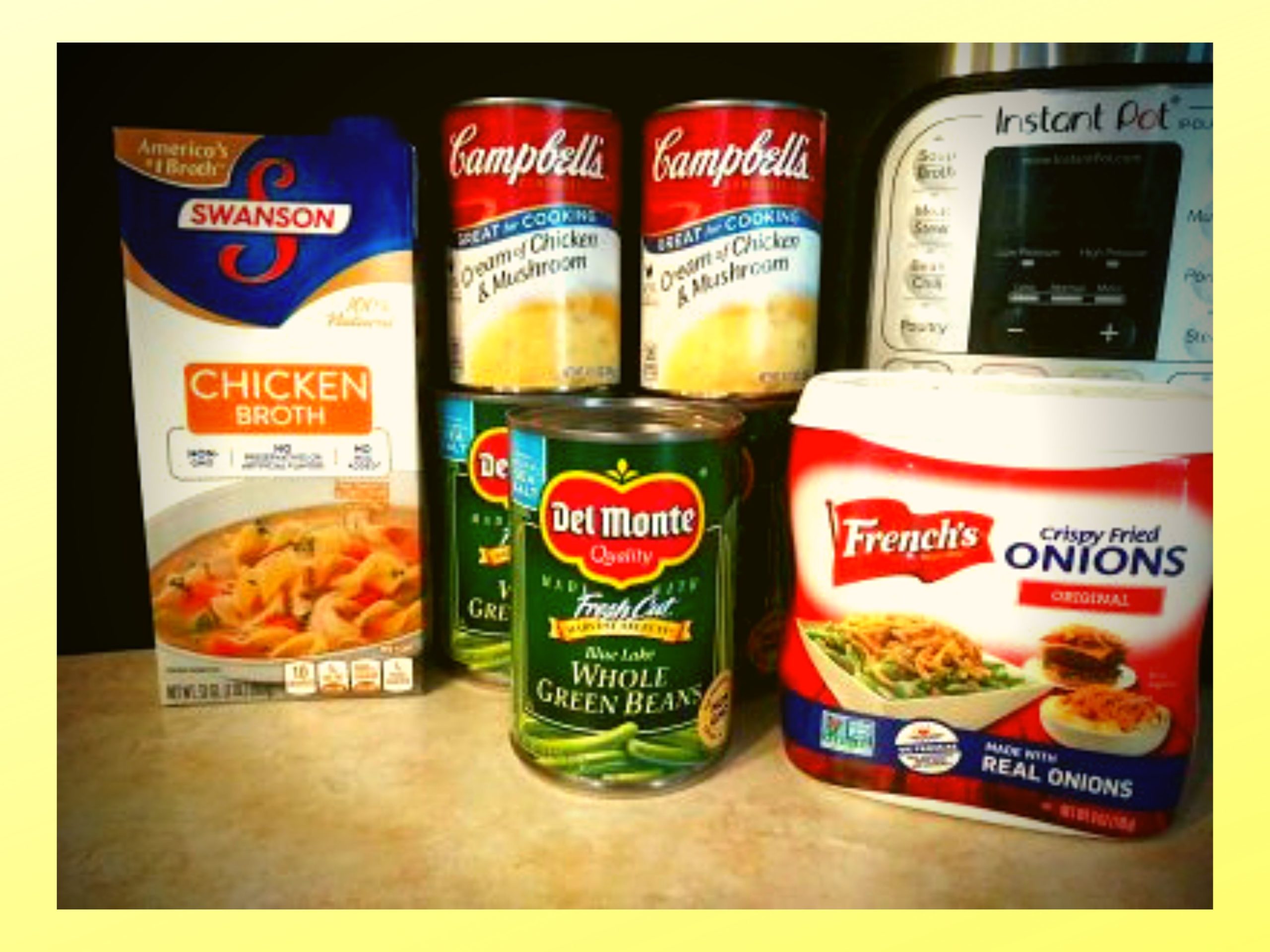 A container of chicken stock, 2 cans of cream of mushroom with cream of chicken, 3 cans of whole green beans, and one container of fried onions sitting on a counter in front of an Instant Pot.