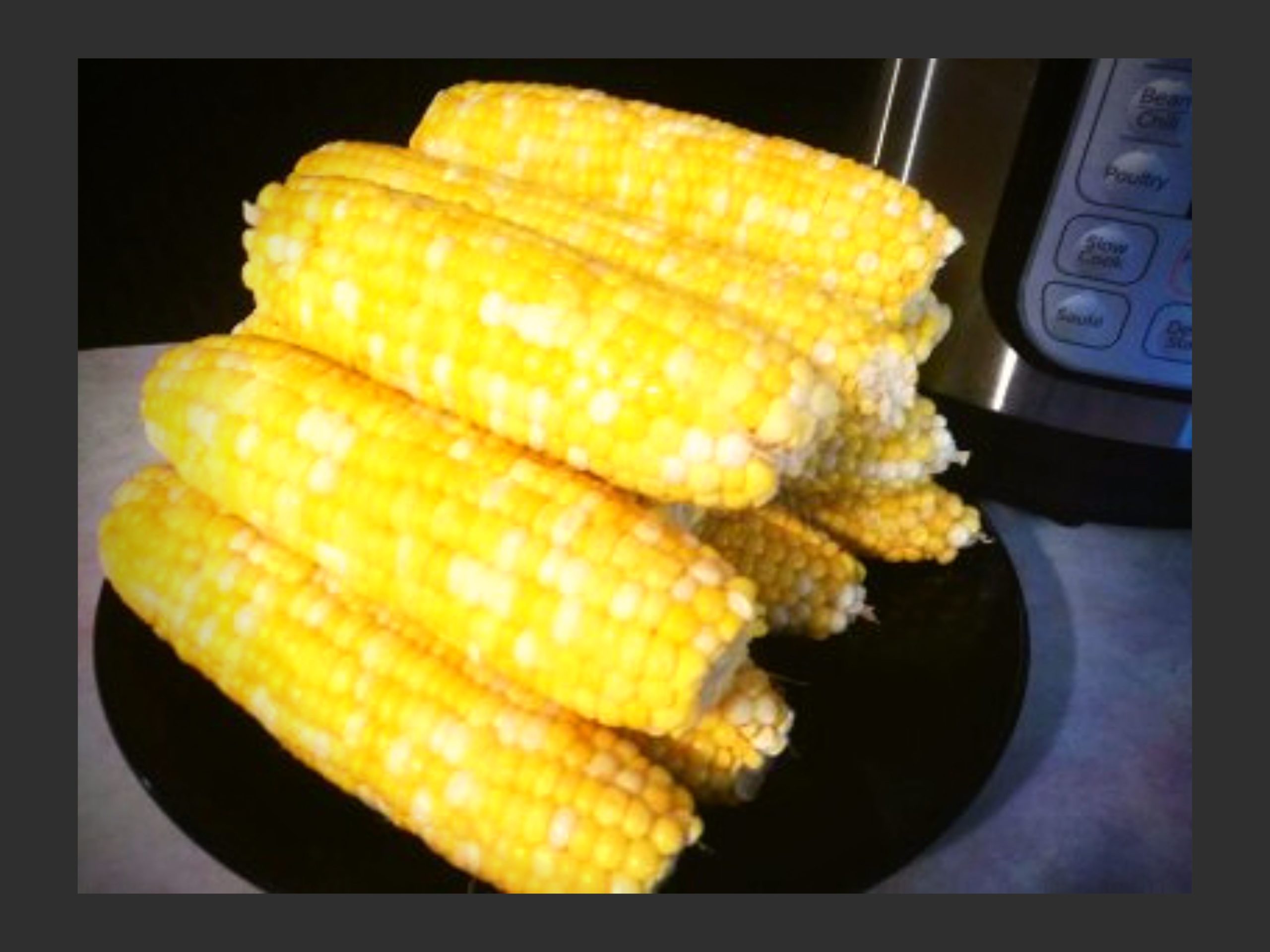 A black plate filled with cooked corn on the cob resting on a counter in front of an Instant Pot