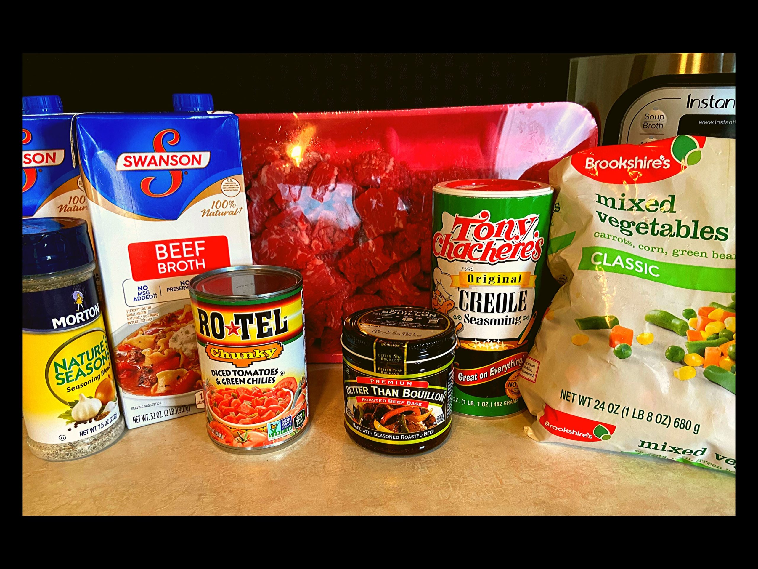 A container of beef broth, can of rotel, raw stew meat, container of Tony Chachere, container of better than beef bouillon, bag of frozen mixed veggies, and an Instant Pot all sitting on top of a kitchen counter.