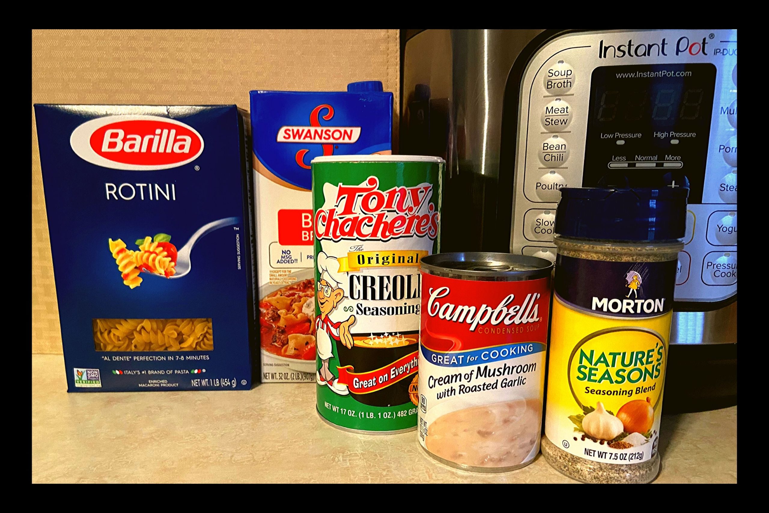 A box of rotini noodles, beef broth, Tony Chachere's Seasoning, Campbells cream of mushroom with roasted garlic, and Nature Seasoning all sitting in front of an Instant Pot on top of a kitchen counter.