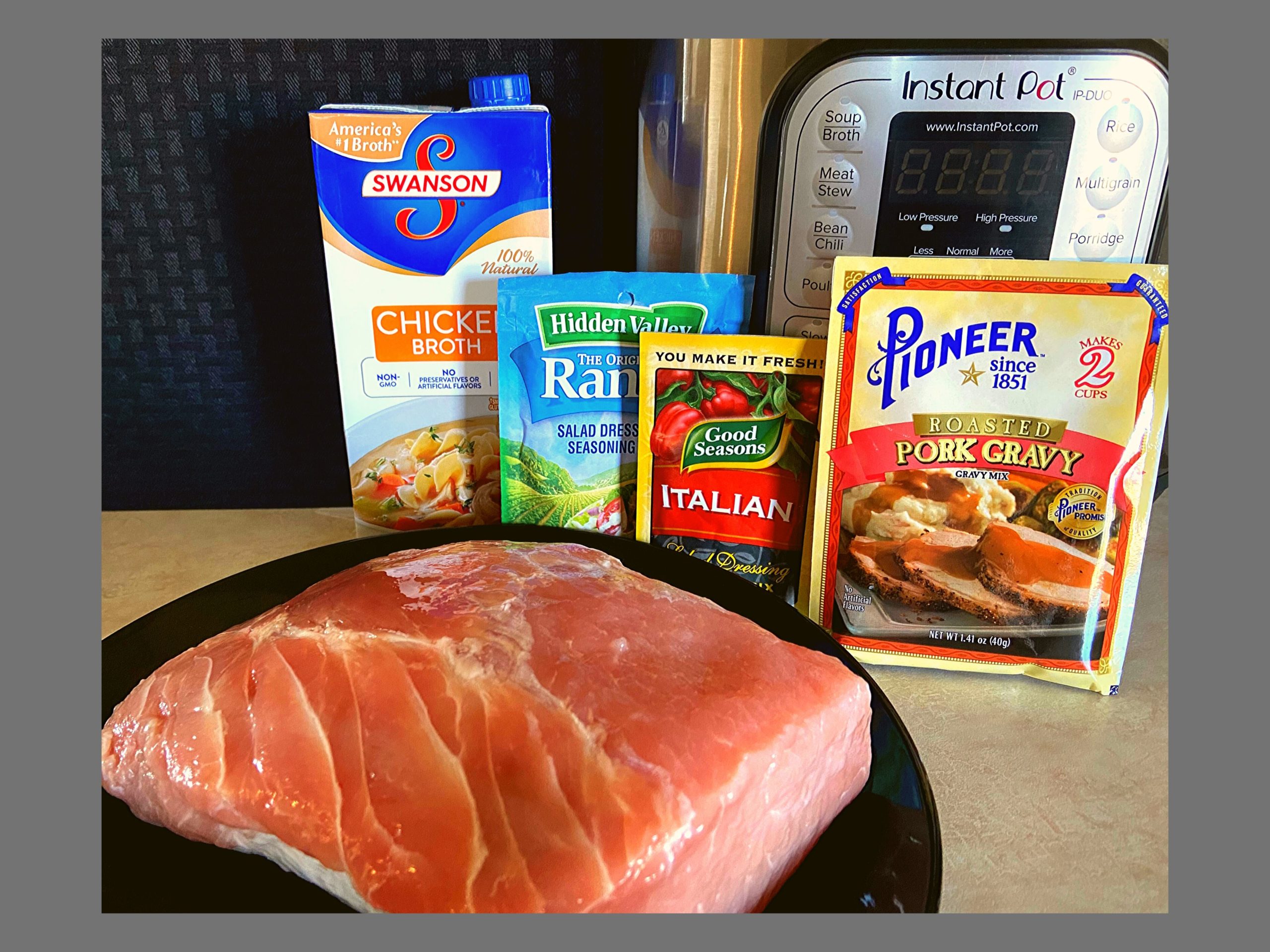 A raw pork roast on a black plate. A container of chicken broth, ranch dressing (dry mix), Italian dressing (dry mix), Pork Gravy (dry mix), and an Instant Pot sitting on a counter top.