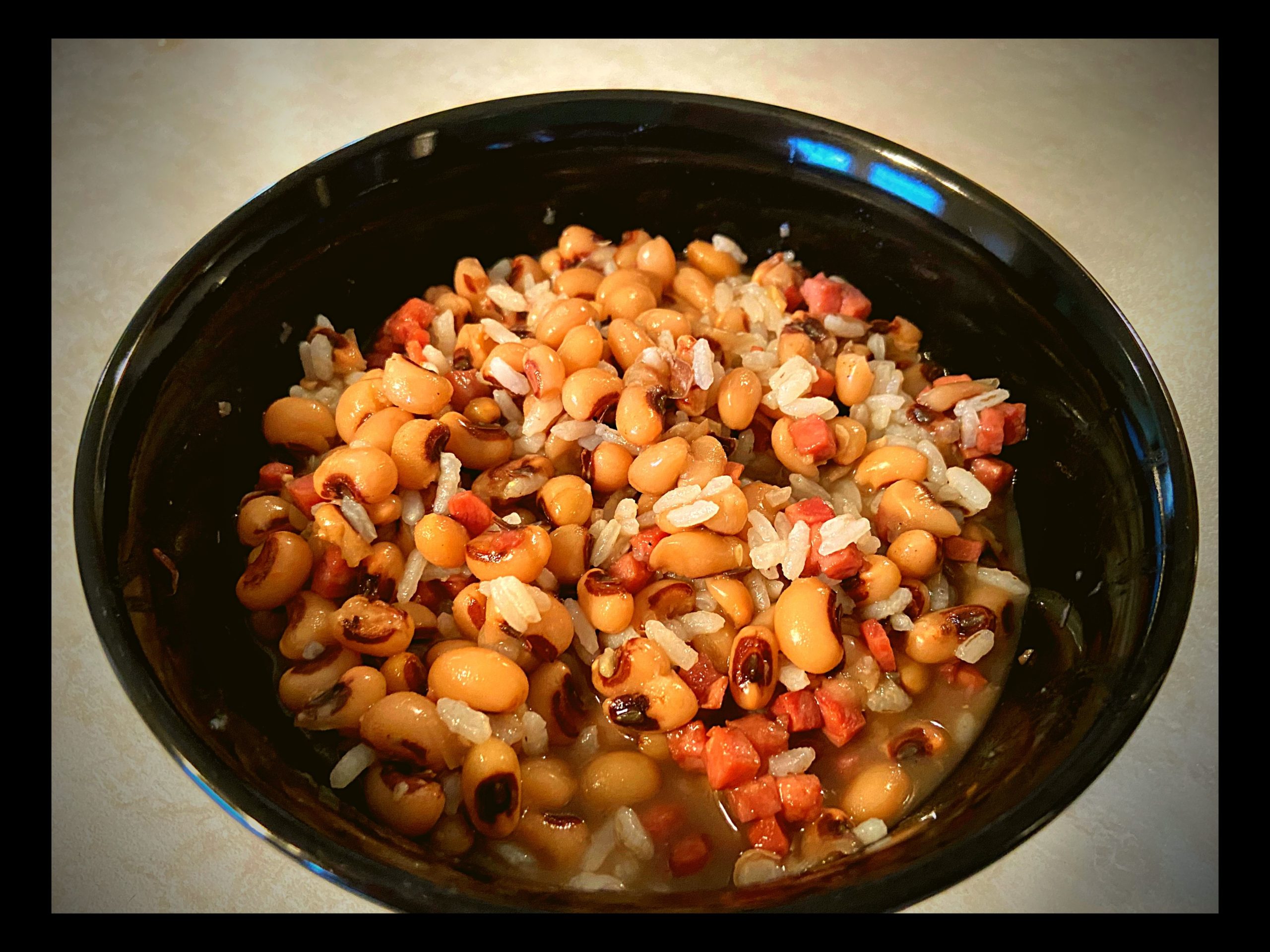 A black bowl filled with black eyed peas and ham mixed with white rice sitting on a counter top.