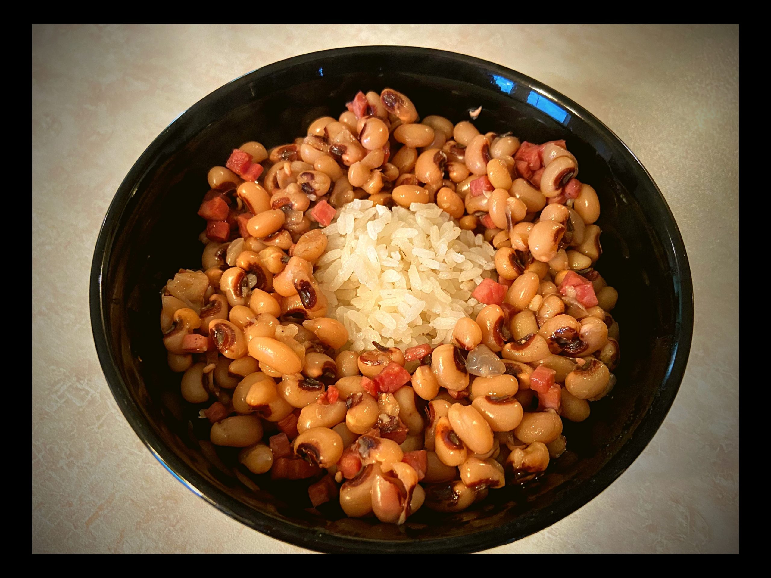 A black bowl filled with a scoop of white rice in the middle surrounded by black eyed peas and ham sitting on a counter top.