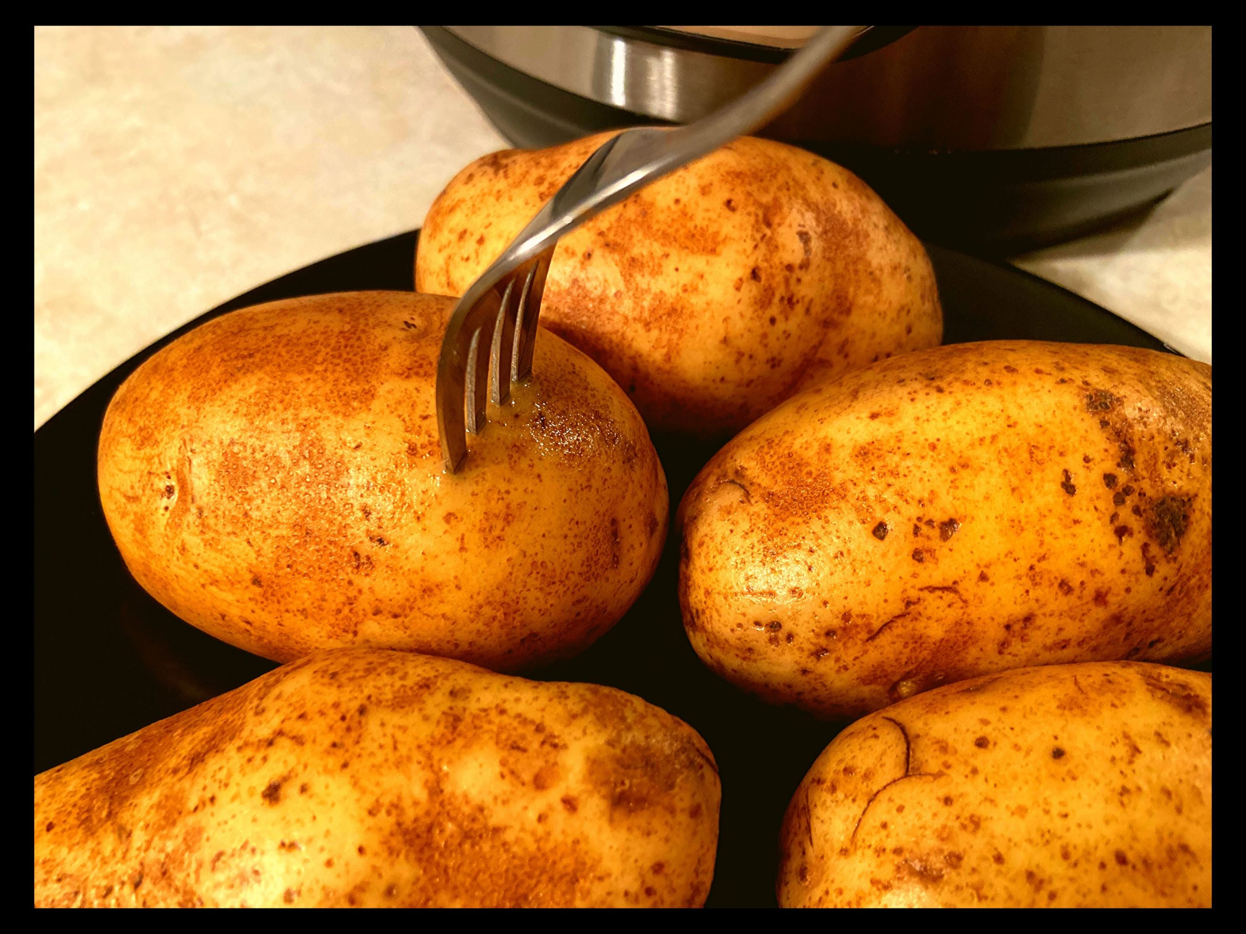 A fork pierced through the side of a raw baked potatoe on a black plate in front of an Instant Pot.