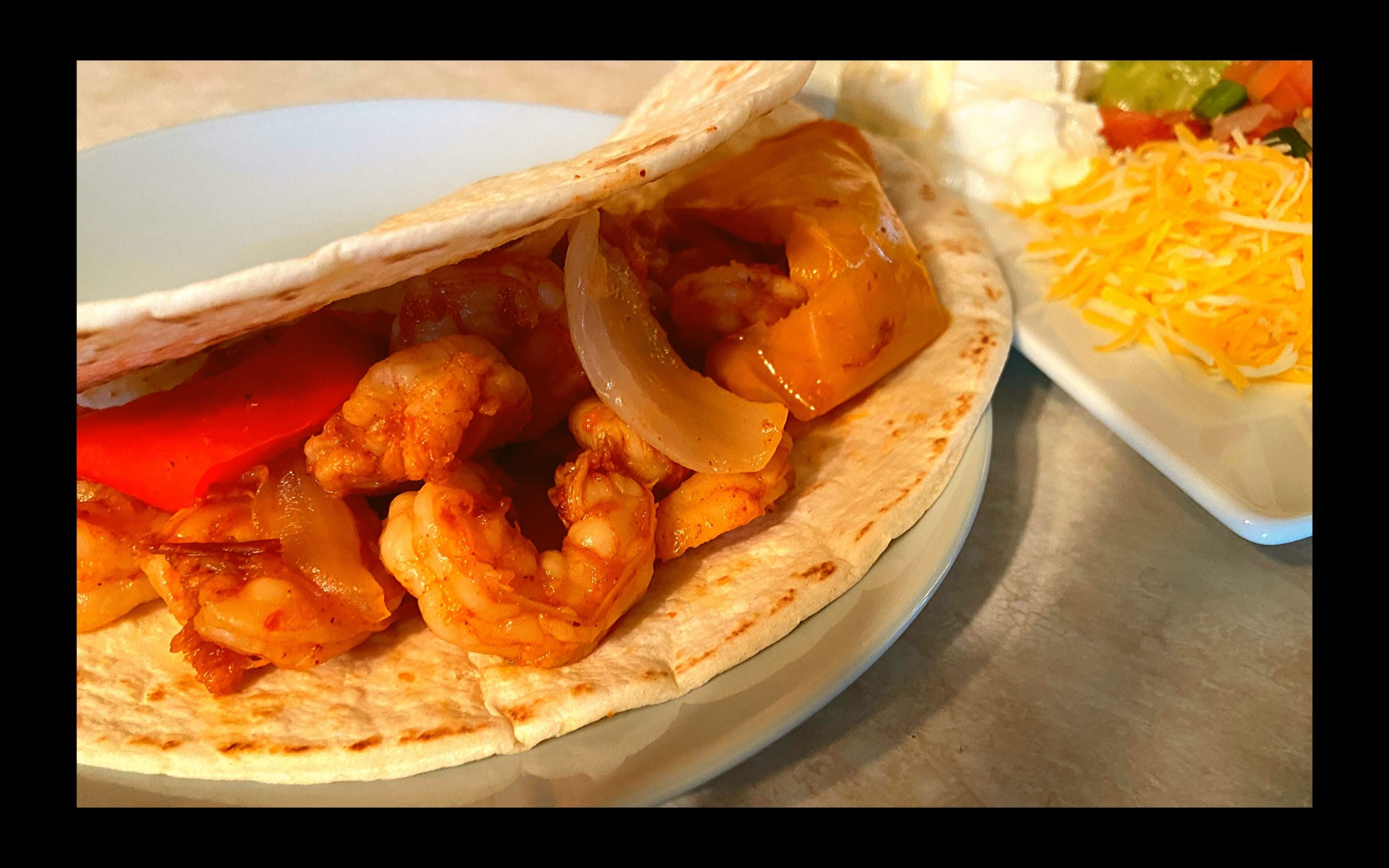 A white plate with a fajita filled with shrimp, onions, and bell peppers. Shredded lettuce, sour cream, and guacamole in the background.