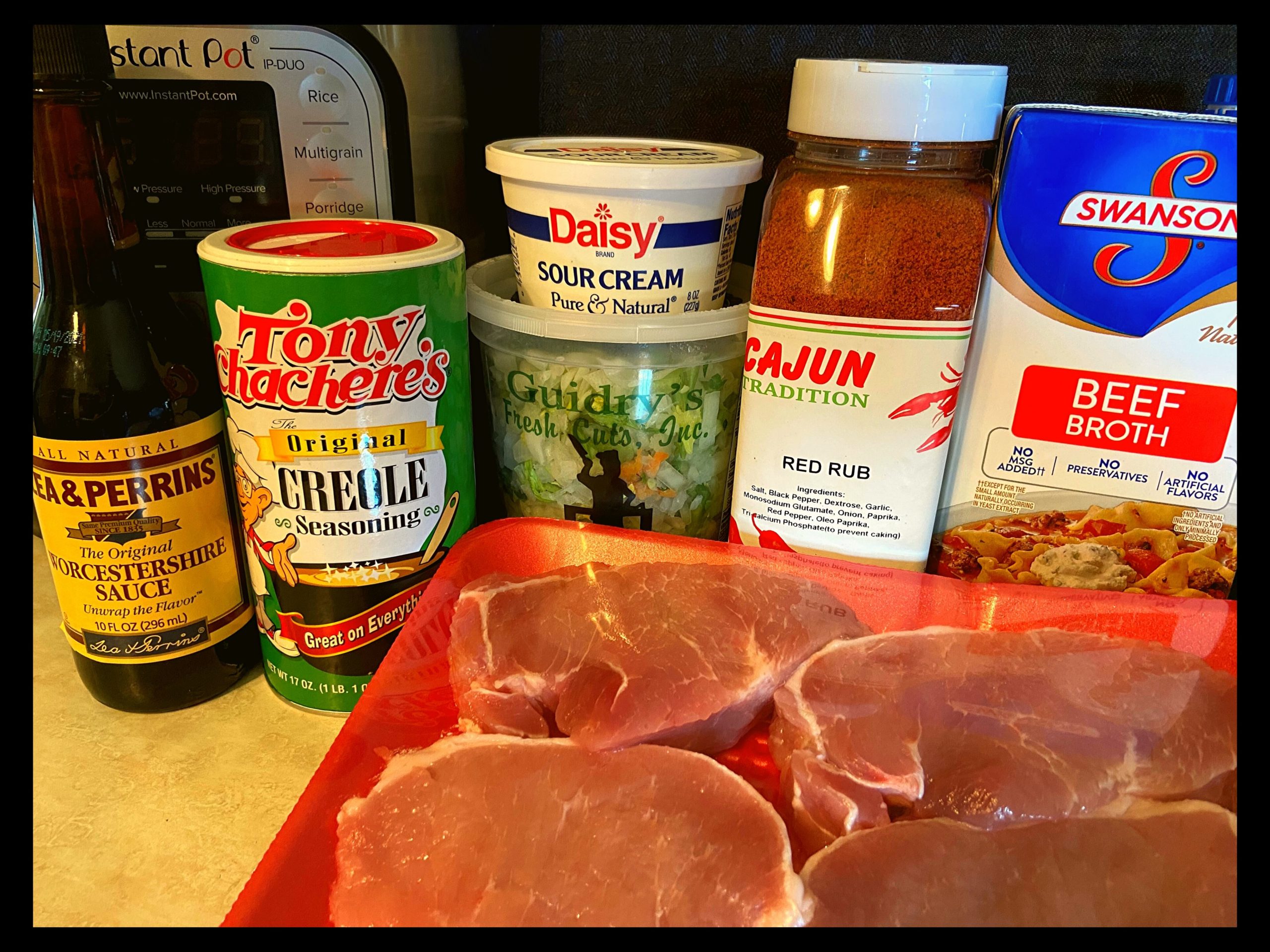 Raw pork chops, worcestershire sauce, tony chachere's seasoning, diced onion, bell pepper, celery, container of sour cream, cajun red rub seasoning, and beef broth on a kitchen counter in front of an Instant Pot.