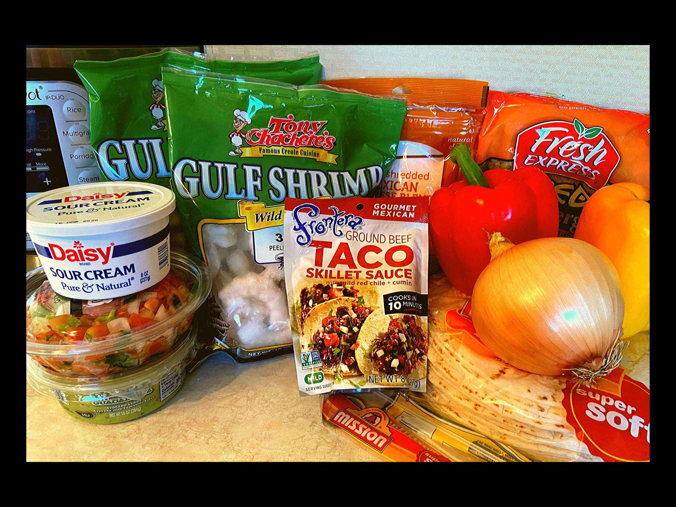 Two bags of large frozen shrimp, container of guacamole, container of sour cream, container of pico de gallo, bag of shredded cheese, pakcket of Frontera Taco Seasoning, red bell pepper, yellow bell pepper, a whole onion, a bag of tortillas, and a bag of shredded lettuce all resting on a kitchen counter top in front of an Instant Pot.l 