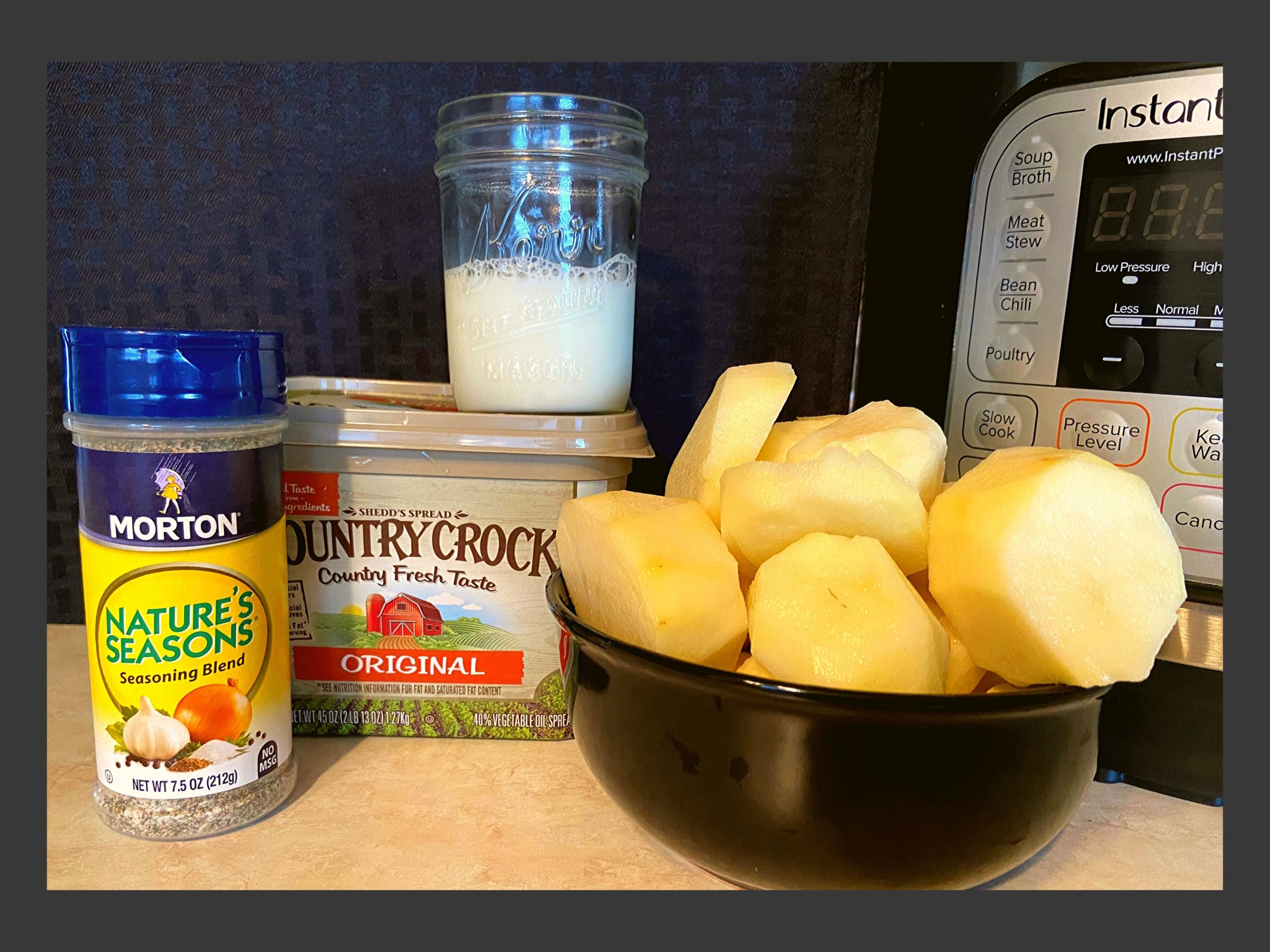 Sliced and peeled potatoes, container of butter, cup of milk, and nature seasoning on a kitchen counter in front of an Instant Pot.