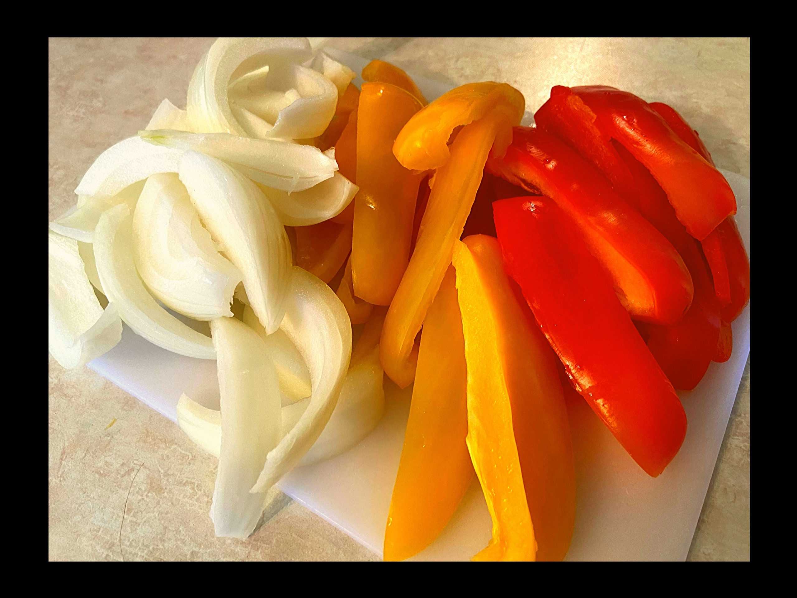 Red bell pepper, yellow bell pepper, and an onion sliced into 1 inch pieces on a white cutting board