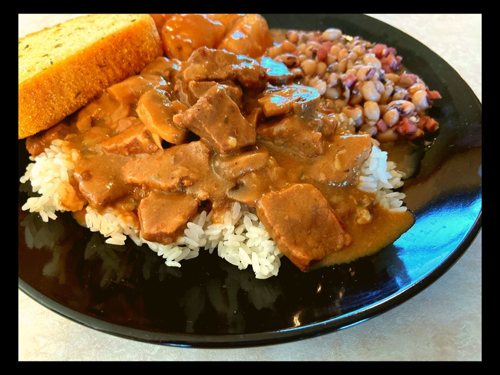 A black plate with garlic bread, black eyed peas, and beef tips with mushroom gravy over rice.