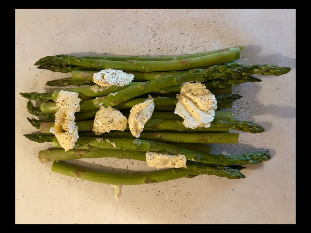 A piece of parchment paper with apsaragus laying on top, sprinkled with nature seasoning and spoonfulls of butter.