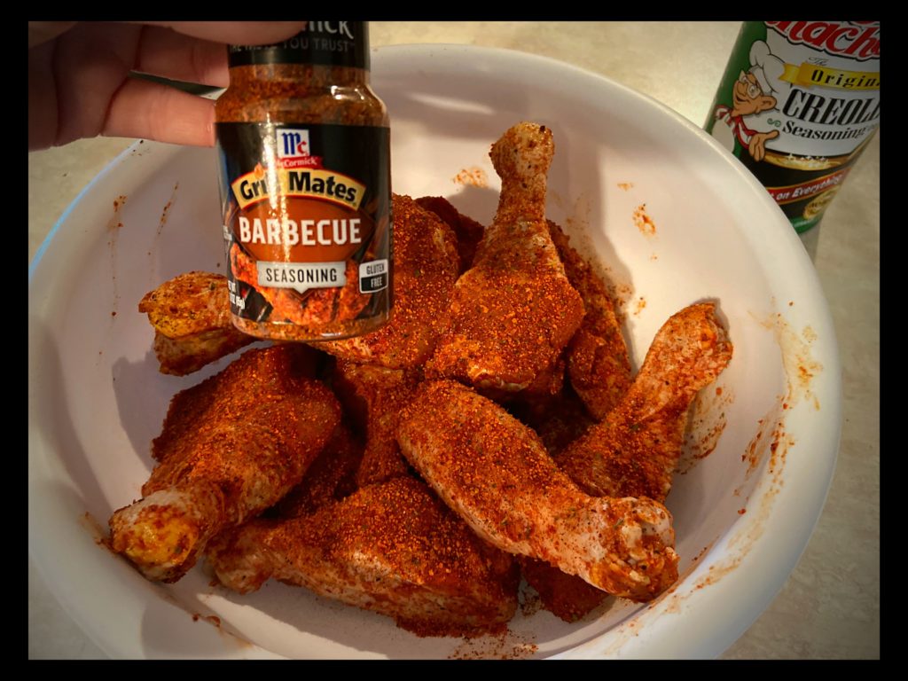 A white bowl filled with raw chicken drumsticks covered in McCormick BBQ Seasoning, Tony chachere's Seasoning, and Cajun Country Red Rub Seasoning.