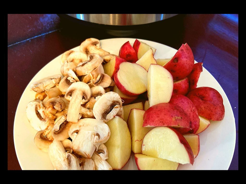 A white plate with sliced white mushrooms and sliced red potatoes sitting on a counter top in front of an Instant Pot