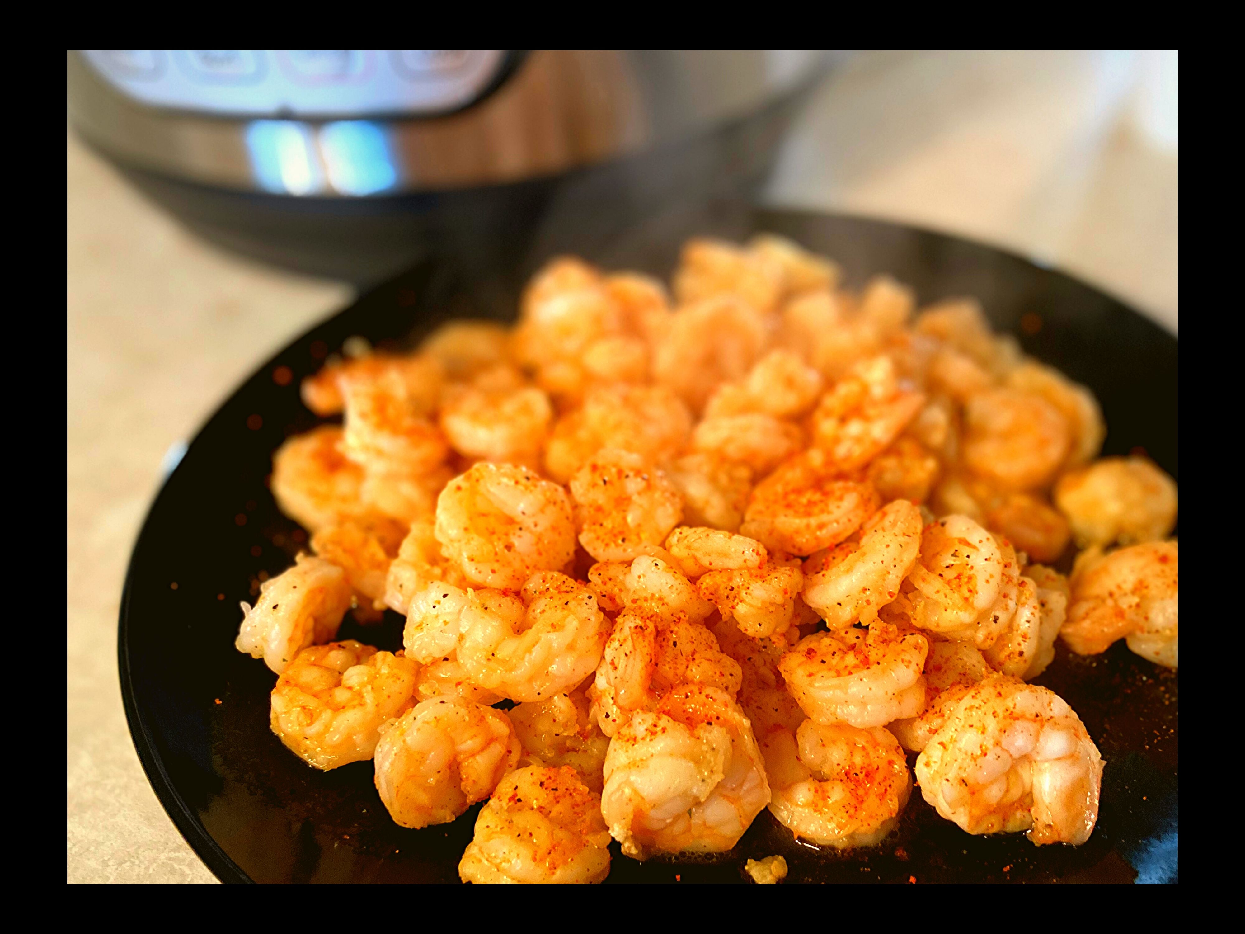 A black plated filled with Easy Instant Pot 3 Ingredient shrimp sitting on a kitchen counter top.