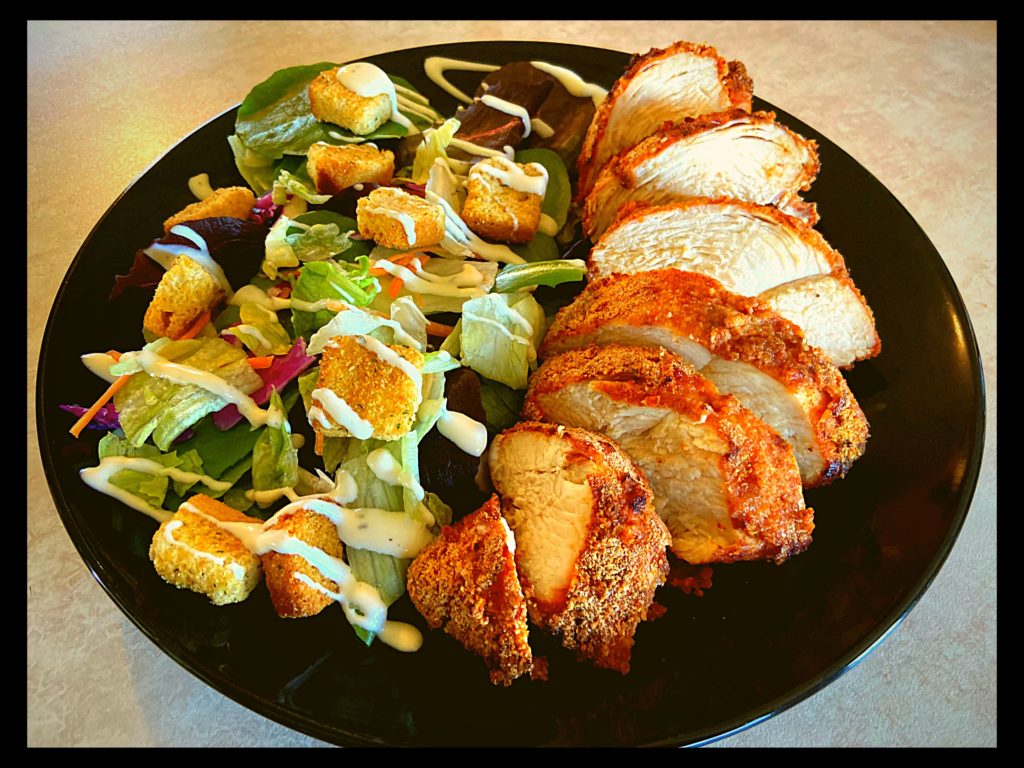 A black plate with a fresh green salad and a breaded air fried chicken breast.