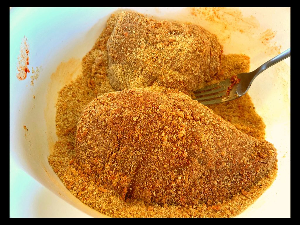 Two raw chicken breasts covered in breadcrumbs in a white bowl.