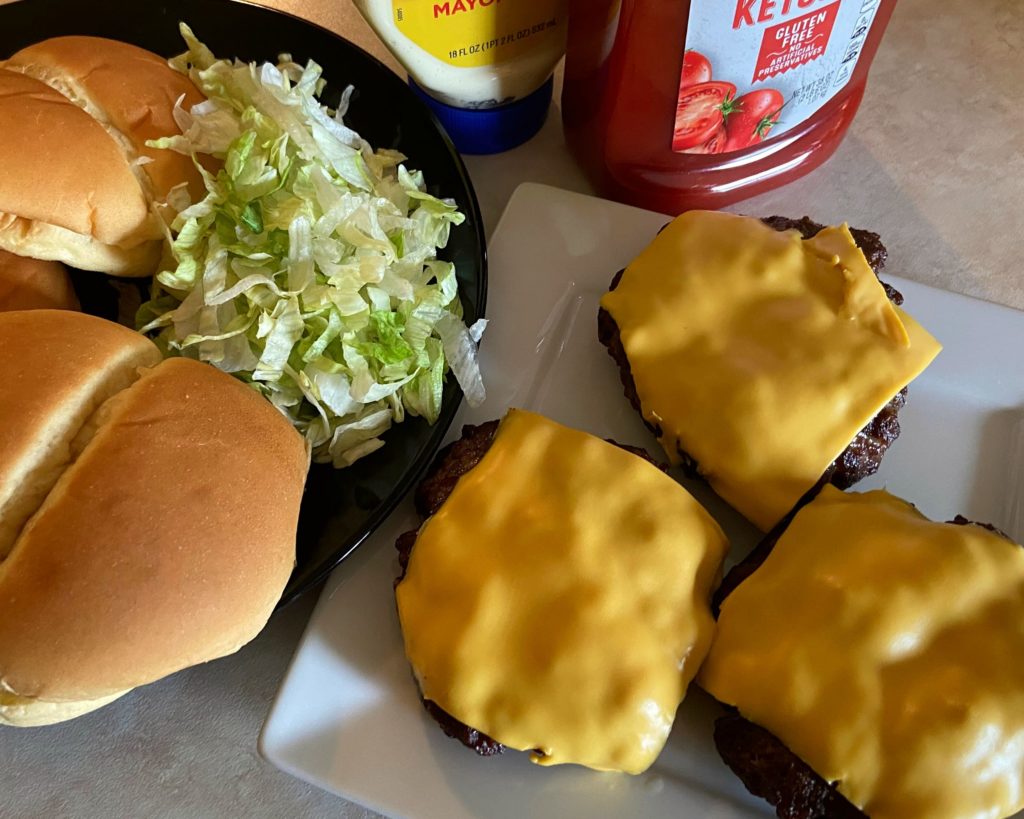 3 cooked hamburger patties on a white plate with a slice of melted cheese on top sitting next to some hamburger buns and lettuce.