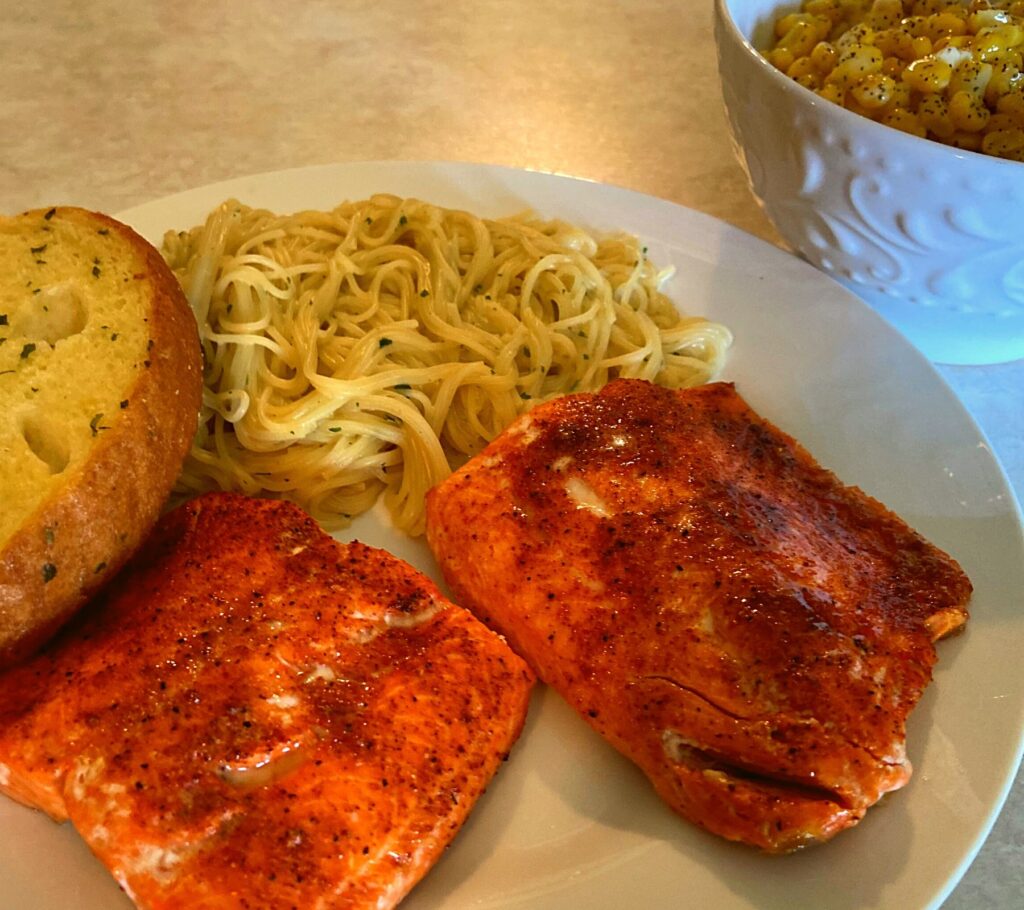 A white plate with Instant Pot Salmon fillets, angel hair pasta, garlic bread, and a bowl of corn.