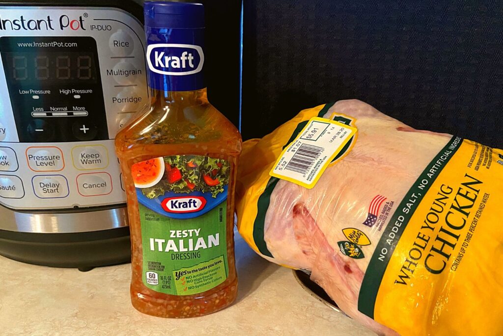 An instant pot, bottle of italian dressing, and a whole raw chicken on a kitchen counter