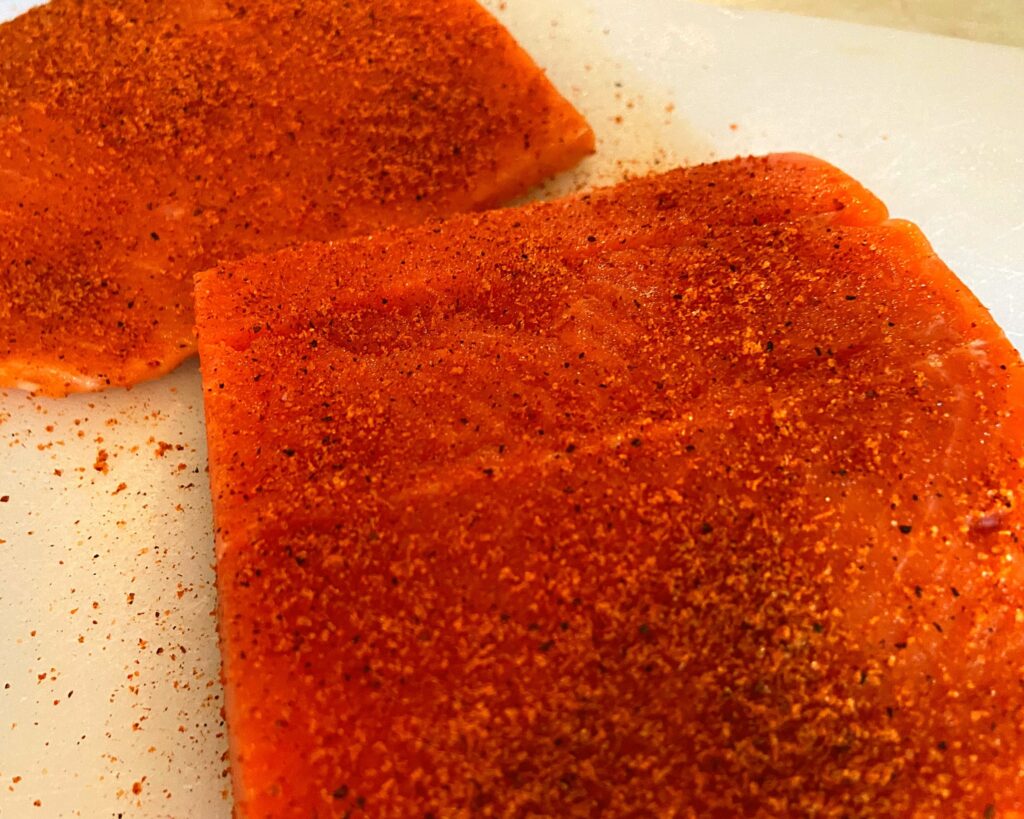 Two raw Salmon fillets on a cutting board sprinkled with tony chachere's seasoning.