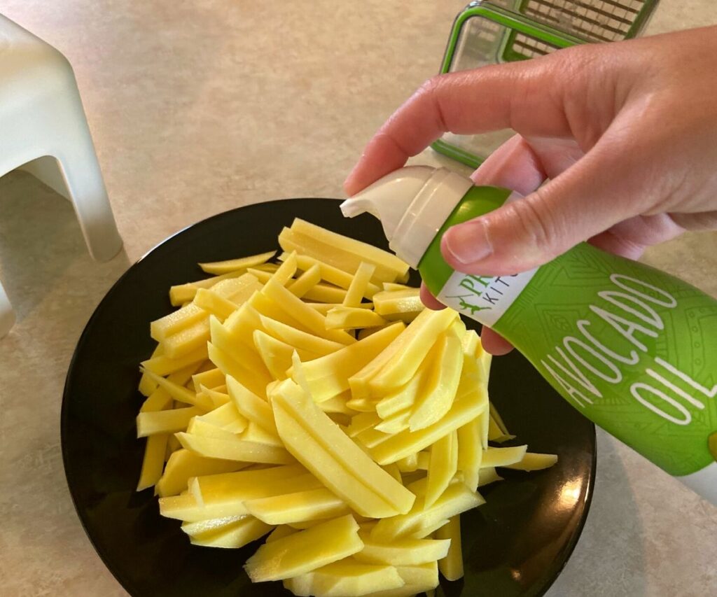 A black plate filled potatoes sliced into french fries being sprayed with Avocado Oil spray.