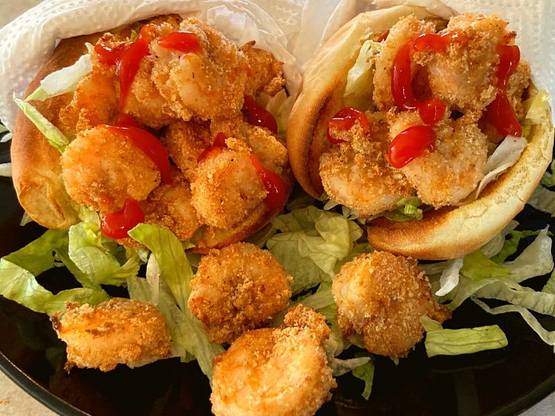 Air fryer fried shrimp in po boy buns with shredded lettuce and ketchup on a black plate.