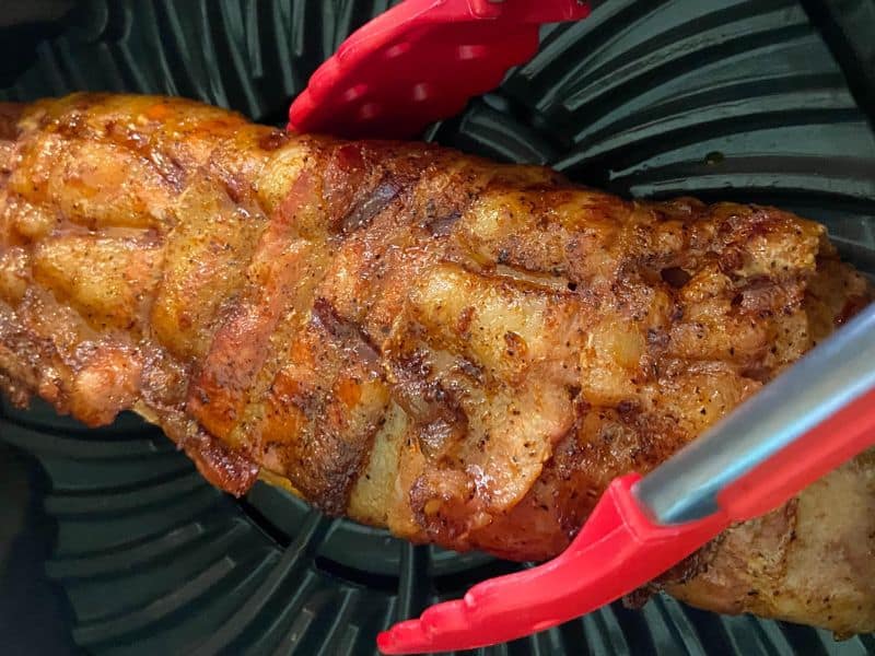 A person picking up a pork tenderloin using cooking tongs from inside of a Ninja Foodi Grill