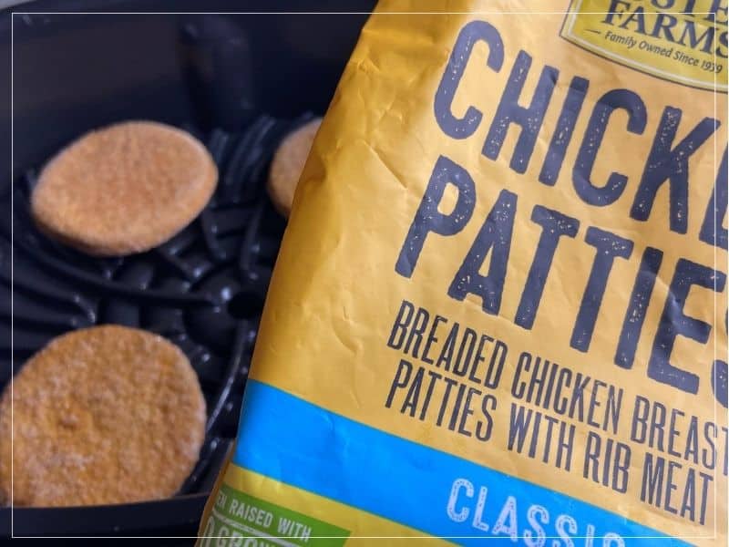 Someone holding a bag of frozen chicken patties over an air fryer with 3 frozen chicken patties sitting in it before cooking.